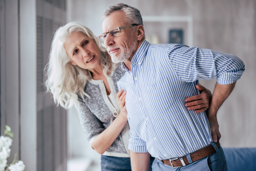 Older man with lower back pain assisted by older woman to stand straight