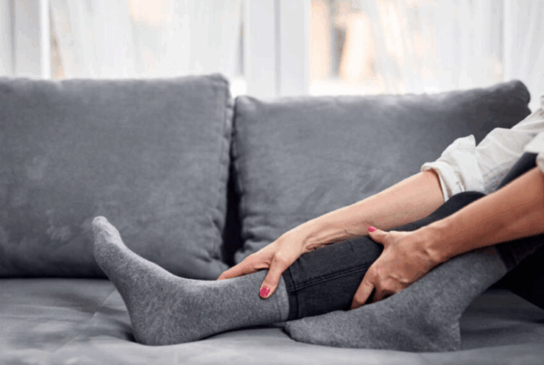 A person sitting on a couch, wearing grey socks and holding their lower leg with both hands as if massaging or soothing discomfort.