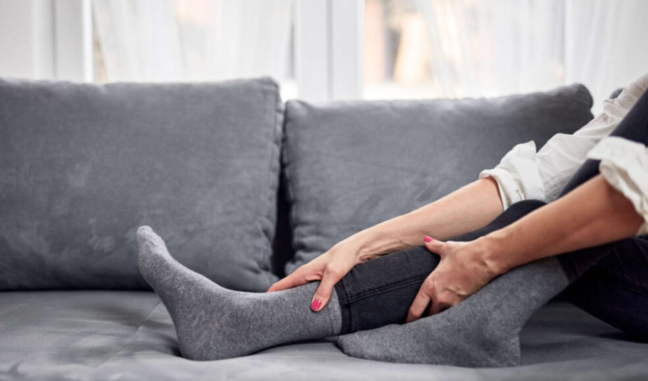 A person sitting on a couch, wearing grey socks and holding their lower leg with both hands as if massaging or soothing discomfort.